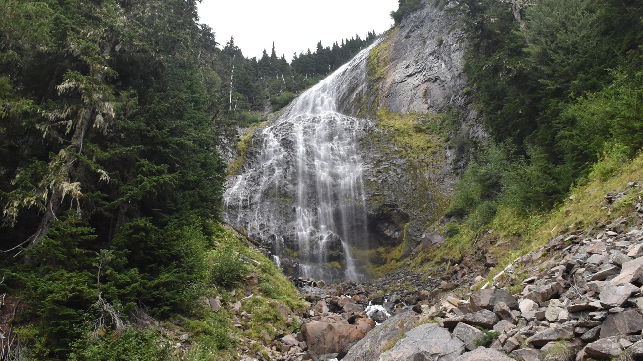 Veil-like waterfall cascades over large cliff with small boulders in foreground