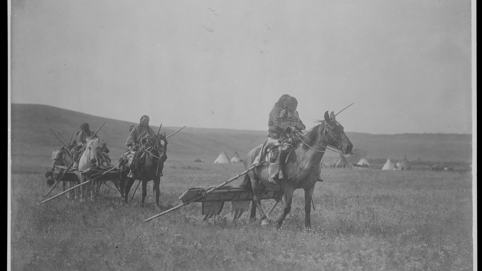 Line of people riding horses on the plains, carrying small loads behind them. Child sitting in front