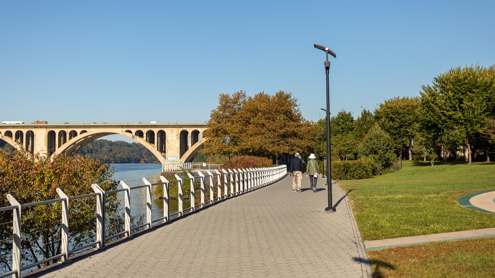 A grassy park next to a trail and river. There is a bridge in the distance.