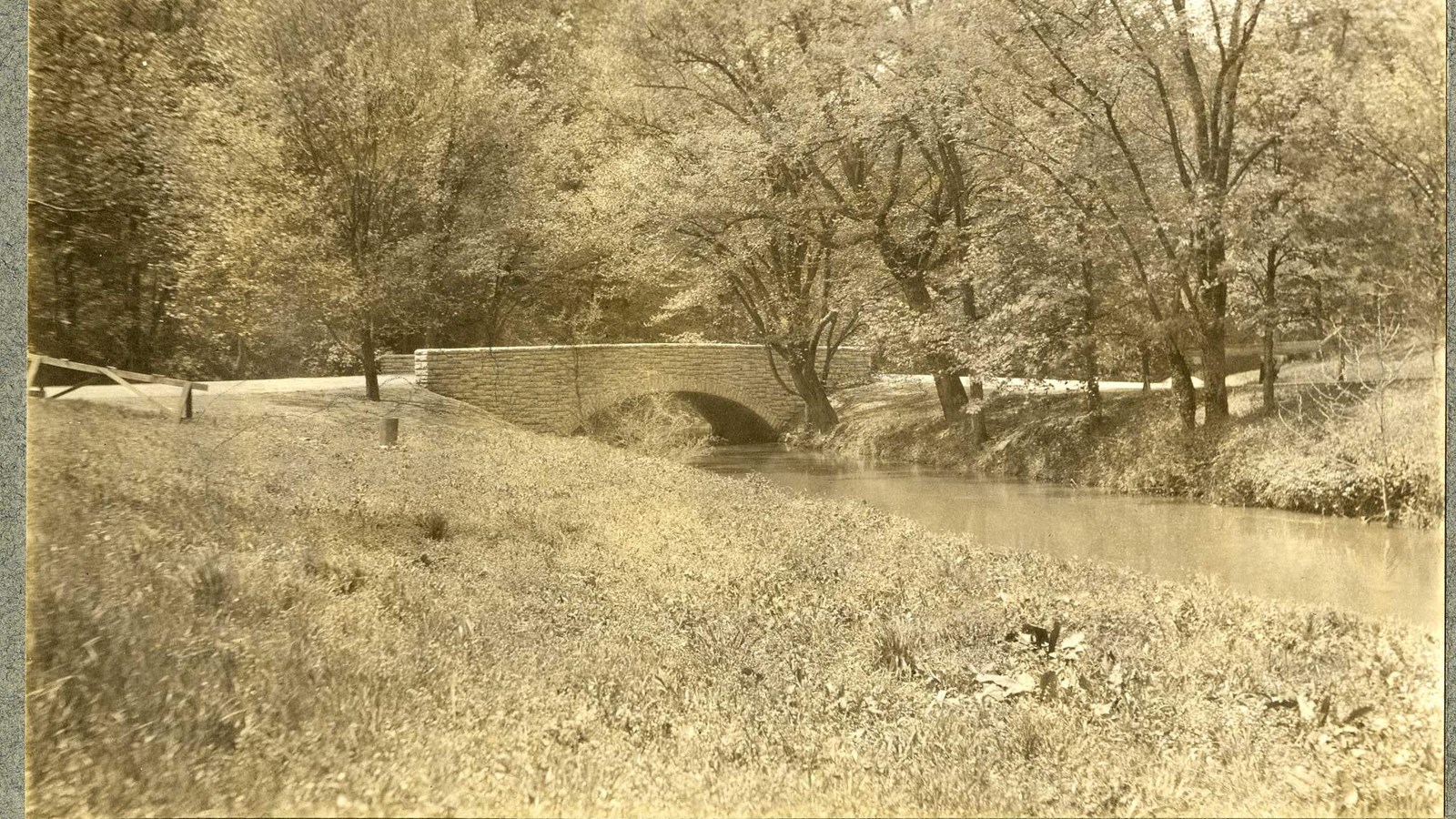 Black and white of grassy area with stone bridge over water with trees by the water