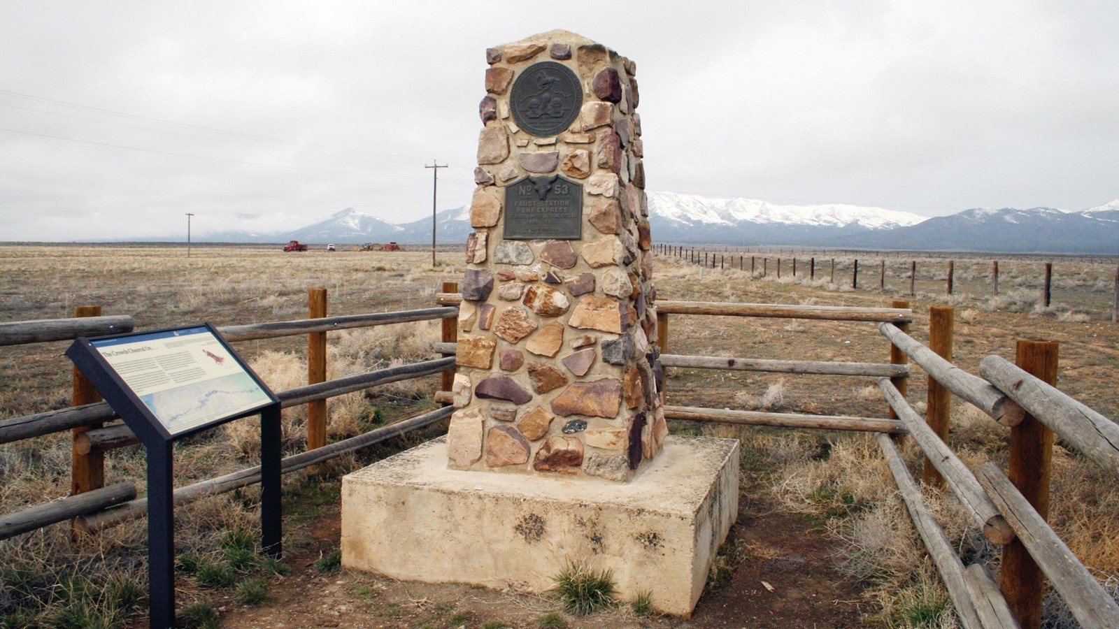 A stone monument four or five feet tall stands next to an upright exhibit panel in a small fence.