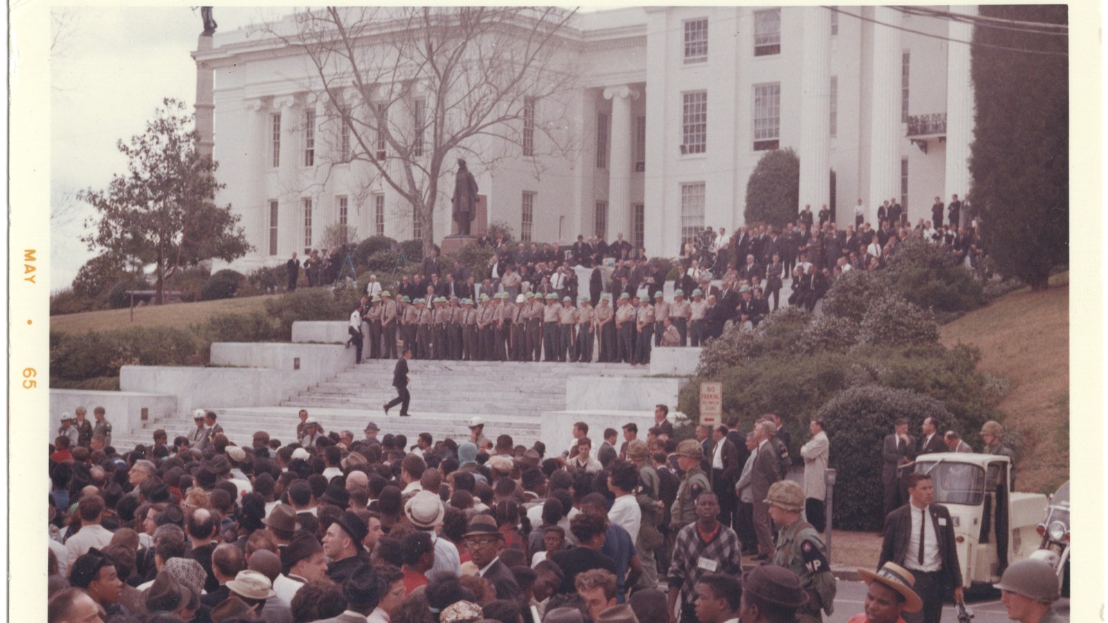 A crowd of people, mostly african american, mull at the bottom of wide white steps before a building