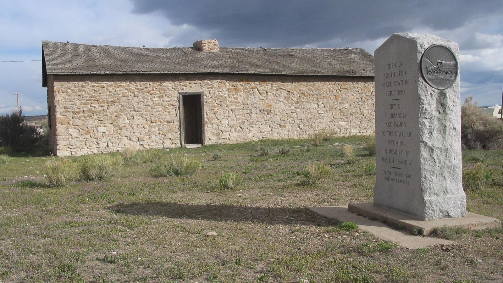 A stone monument stands next to a one story, stone building with a single door.