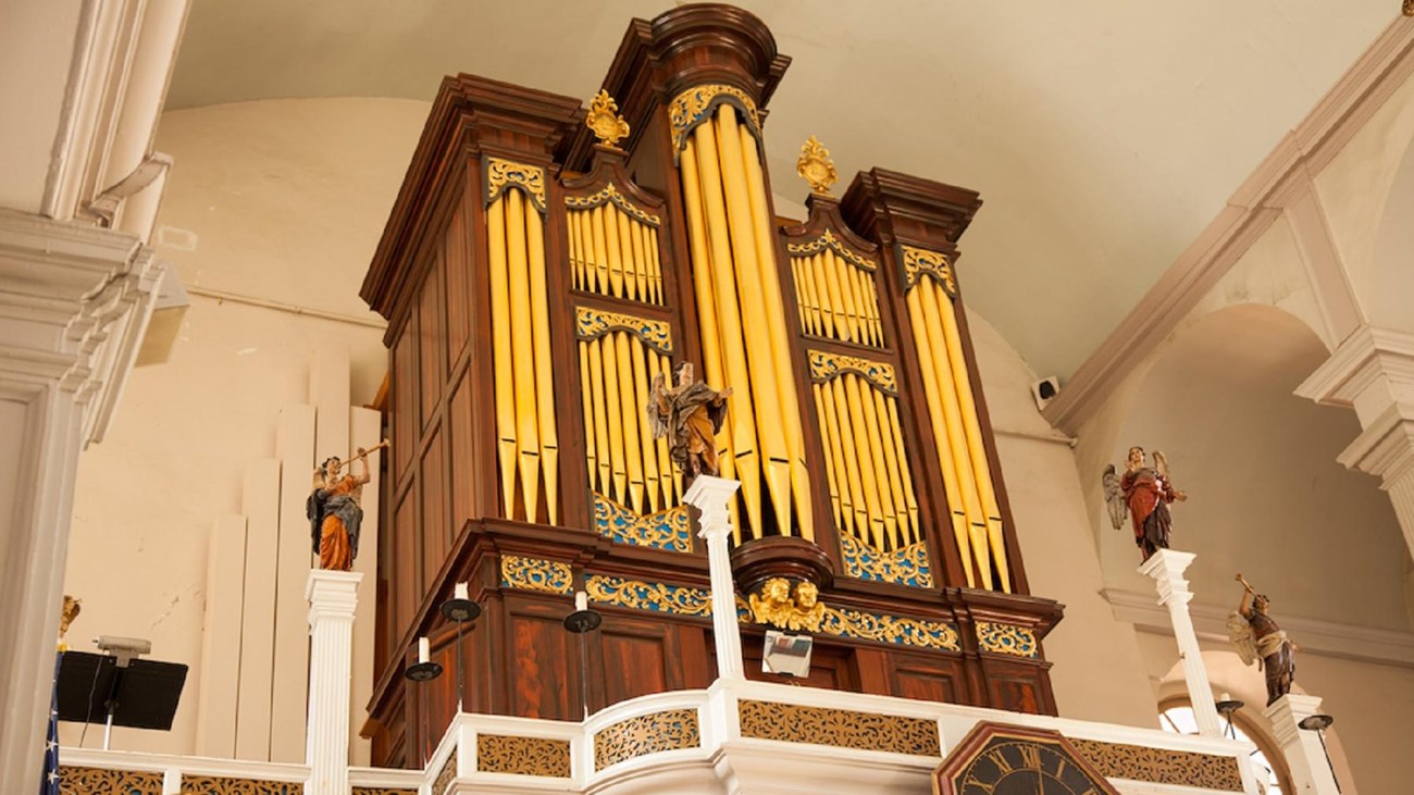 large organ with golden pipes on the second gallery floor of a church. Statues of angels flank it.