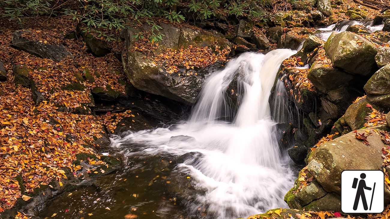 A short, cascading waterfall framed by boulders and orange, fallen leaves. Hiker icon in corner.