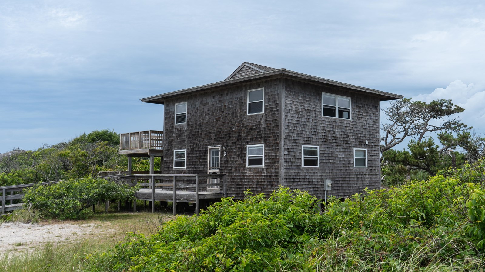 A two-story house with wooden shingles among low-growing beach plants in a sandy area.