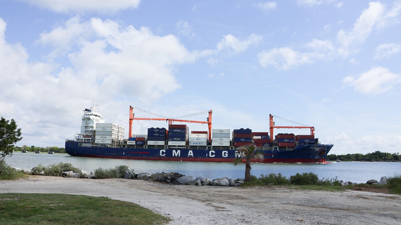 A large cargo ship on a river with a gravel driveway on land.