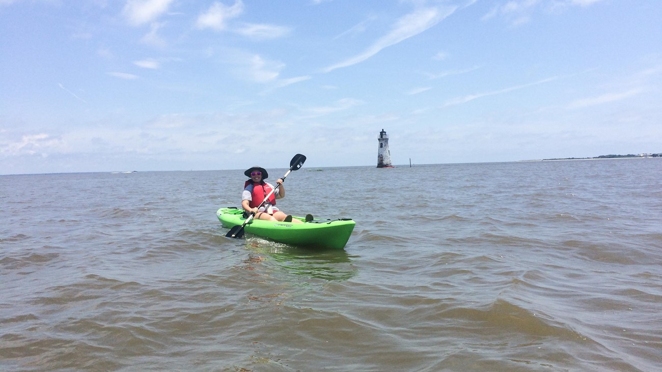 A woman in a green kayak with a lighthouse distantly behind. 