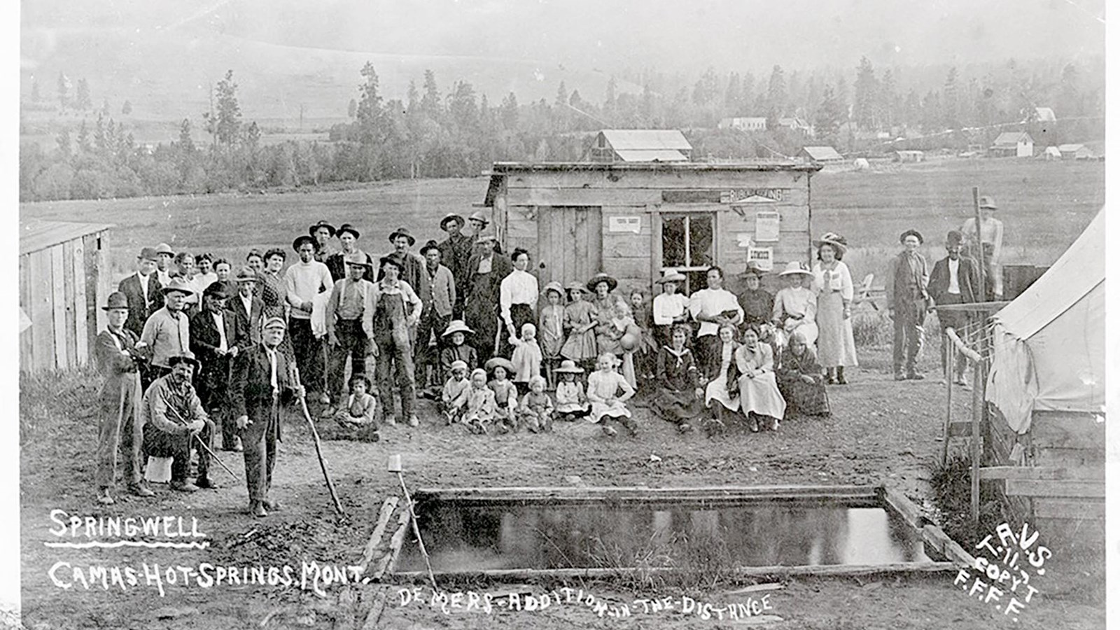 Photograph of many individuals standing in front of a small hut. A hot spring is in the foreground.