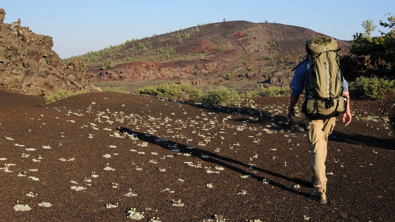 A hiker with a large backpack walks on a cinder trail toward a large volcanic cone.
