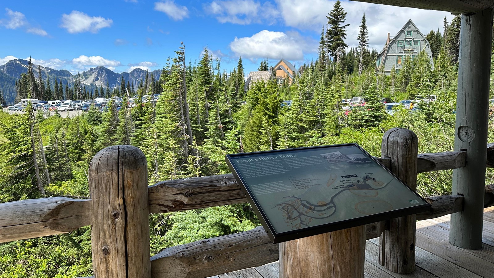 An exhibit panel on a porch with a log fence overlooking meadow, parking lot, and buildings.