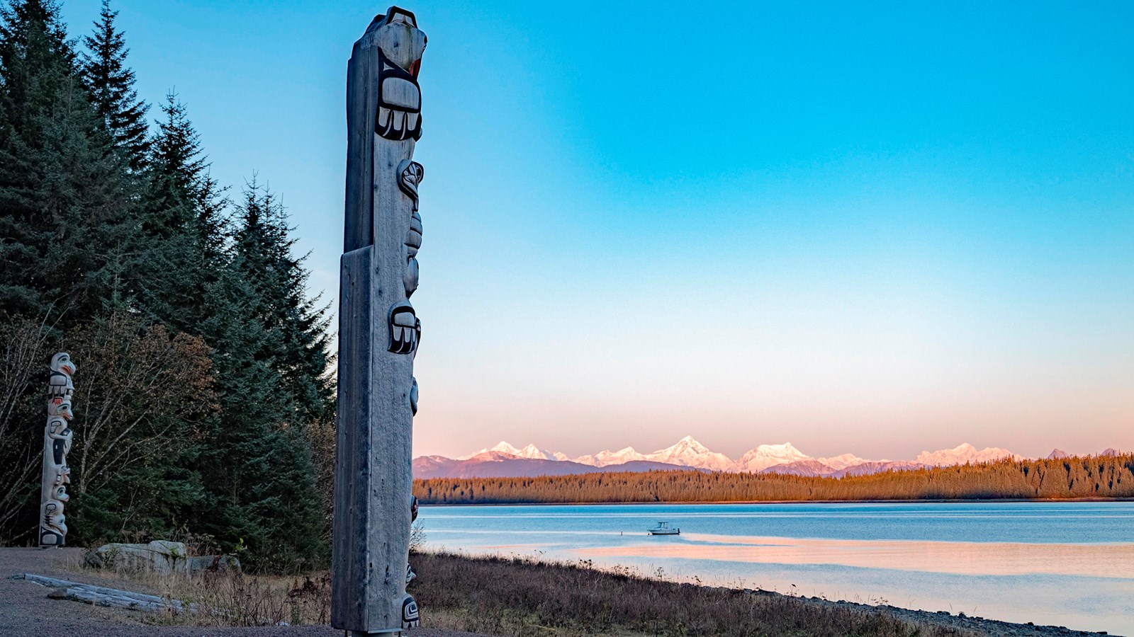 Two totem poles stand pointed toward water. Orange mountains shine in the morning light.