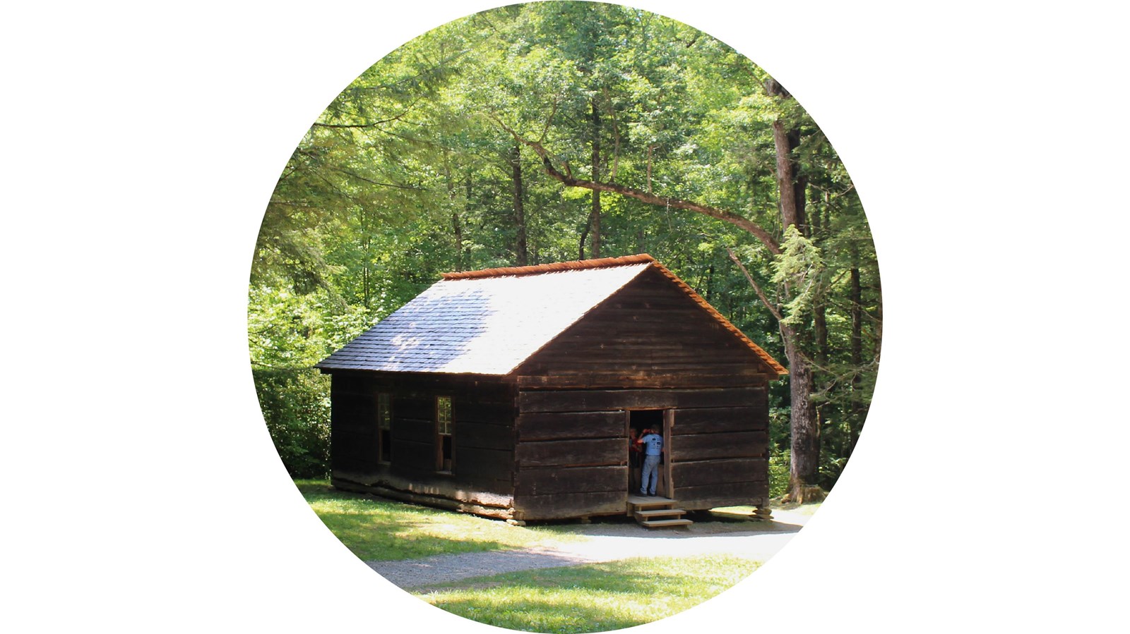 A brown, wooden schoolhouse with a gravel path in front of it. Trees in the background.