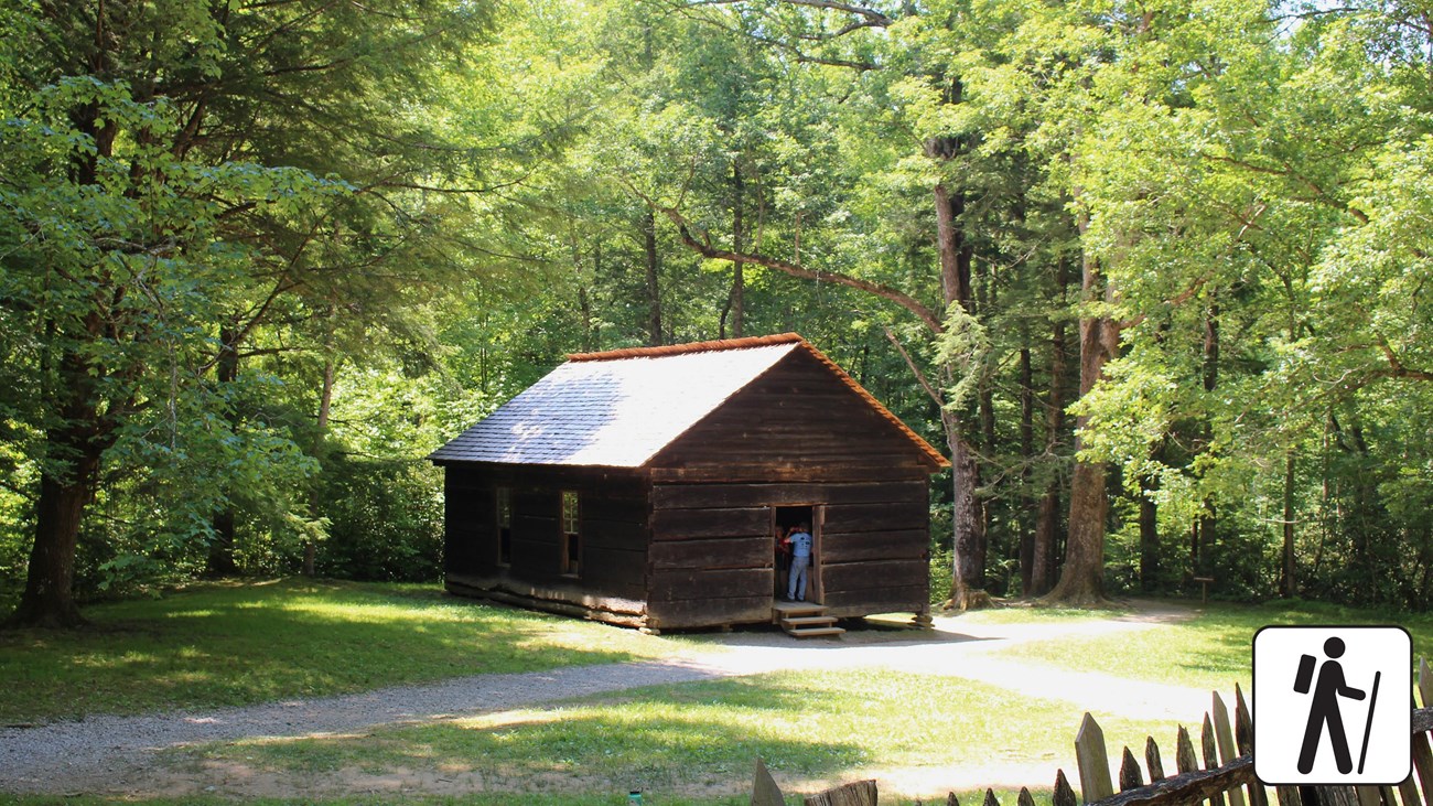 A dark brown, wooden schoolhouse gravel path in front of it. Hiker icon in photo corner.