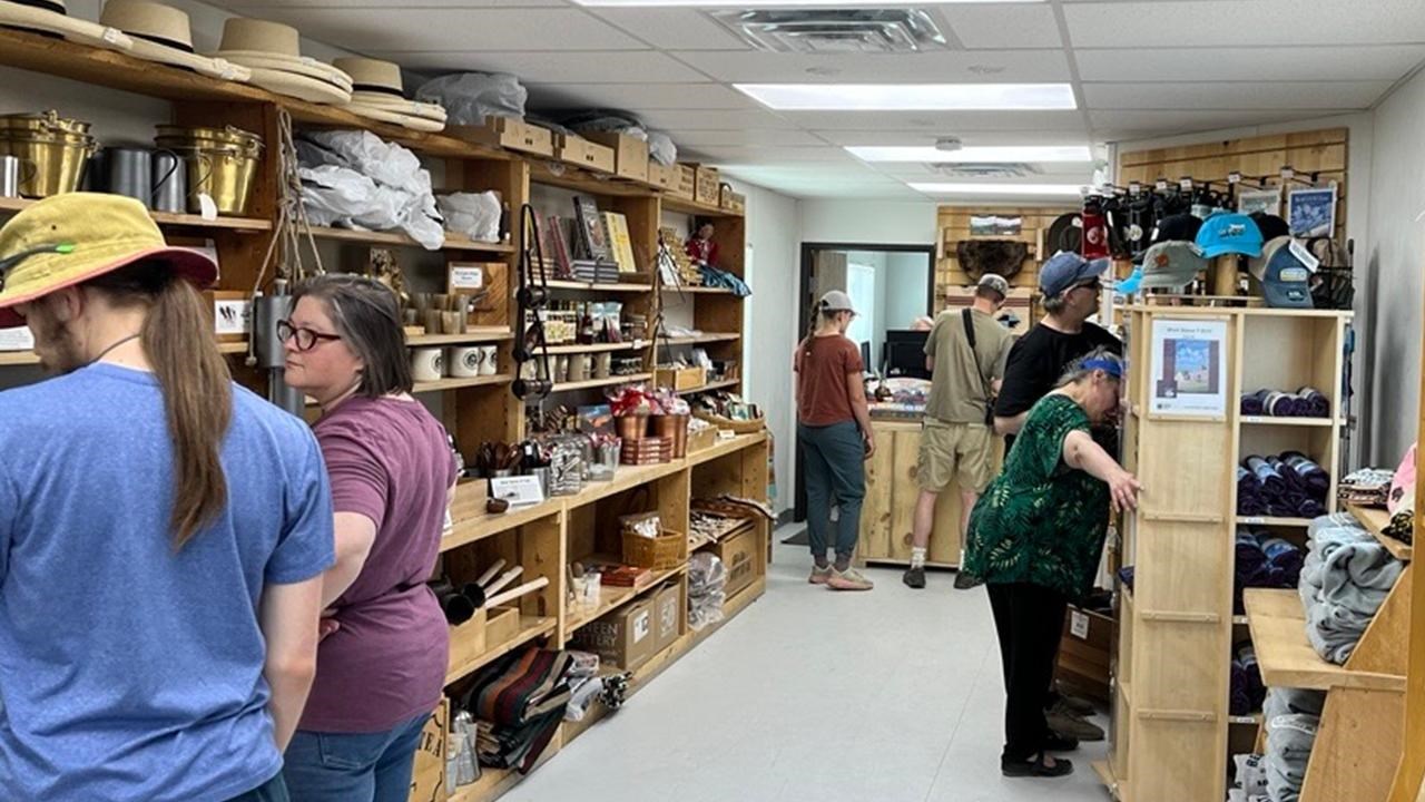 Visitors stand inside a narrow building and review items on wooden shelves. 