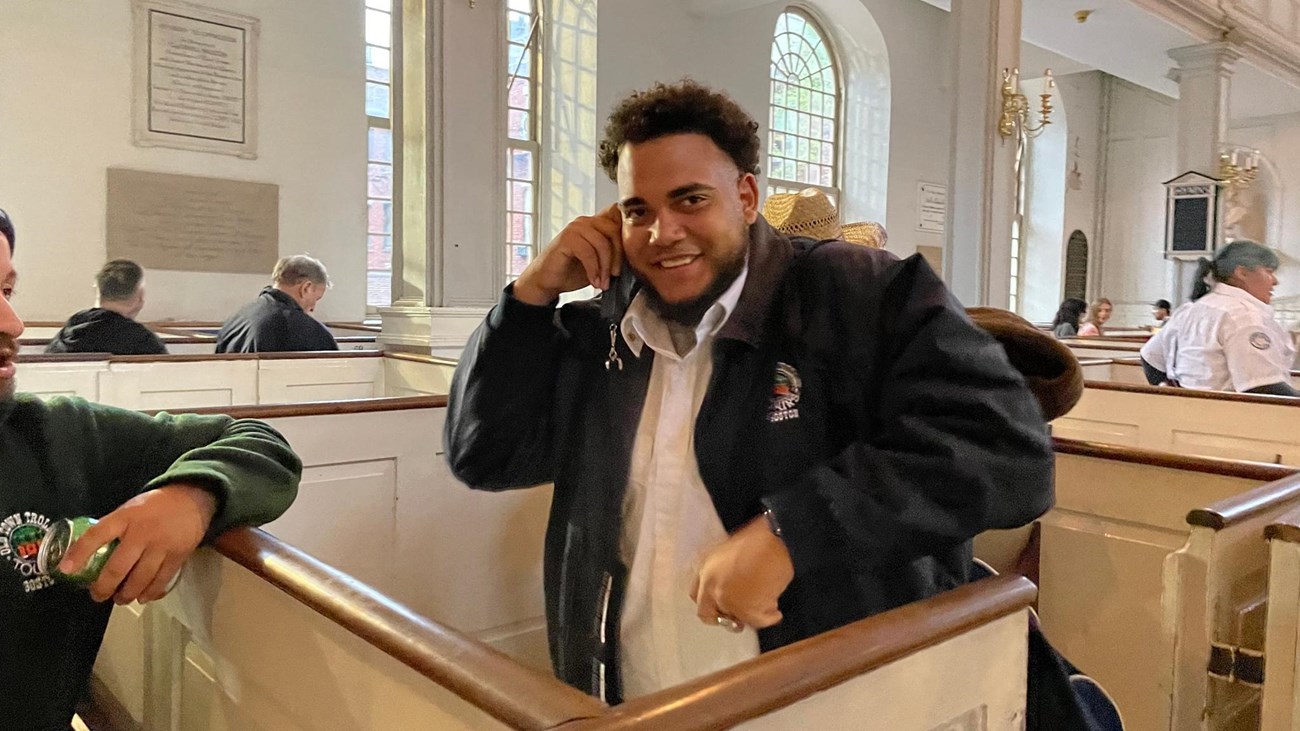 a man holding up an audio tour set to his ear while standing in a pew. 