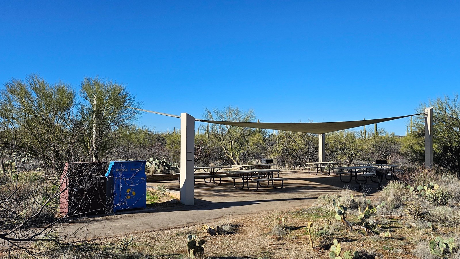 A large triangular shade structure covers six metal picnic tables. Two trash cans left of tables