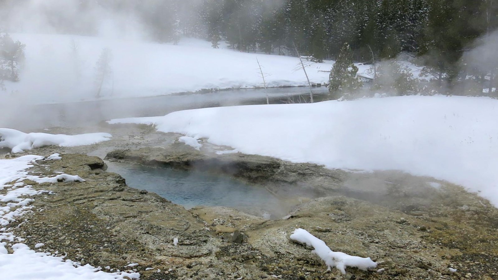 A hot pool with steam above, snow around the periphery, and a river in the background