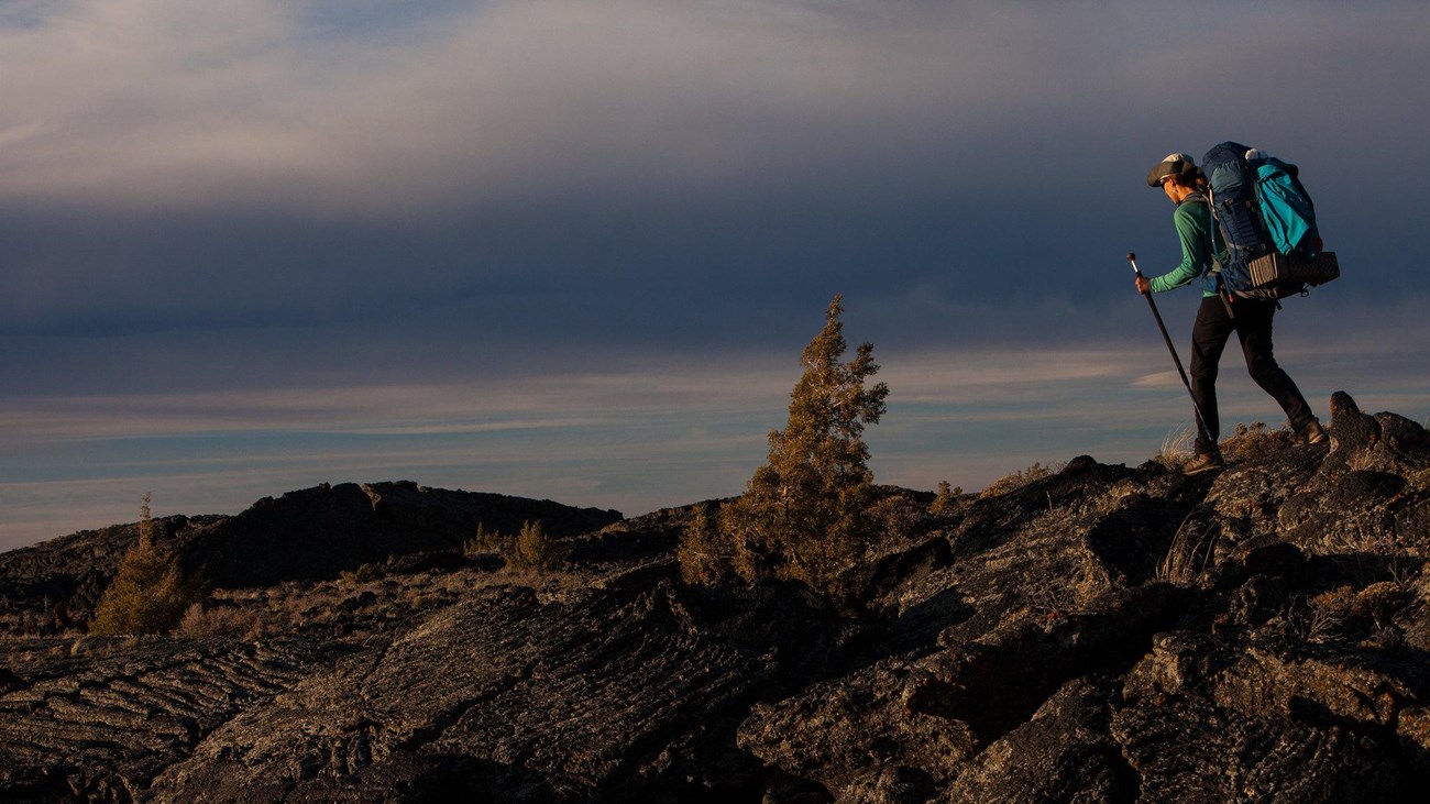 A hiker with a large backpack traverses a lava field.