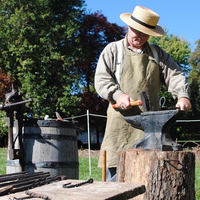 A man with a straw hat bends over a black iron anvil with a hammer. 