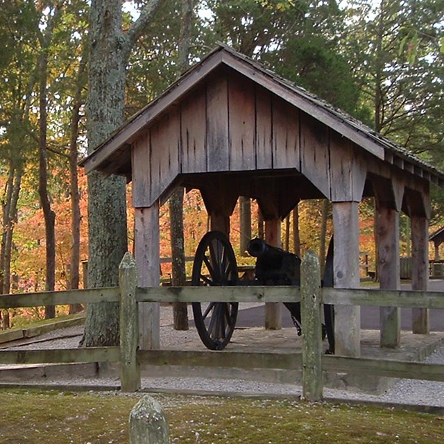 A field artillery gun sits in a covered, wooden pavilion in an autumn-colored woodland.