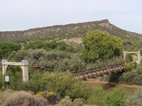 An old, rustic wooden bridge spans a muddy dirty river with a mesa visible in the background.