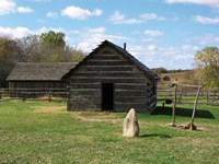 Two log cabin buildings with pitched roofs sitting in a grassy area with trees in the distance.