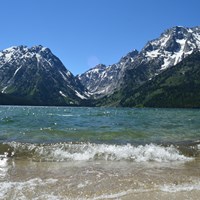 Waves crash on the shore of a clear, blue lake at the base of a mountain range.