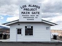 A small white sided building with a sign reading "Los Alamos Project Main Gate" under a cloudy sky