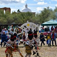apache crown dancers with large head pieces dance with audience watching and church in background