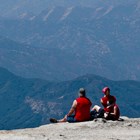 Two adults and a child sit at the edge of a cliff overlooking mountain vistas