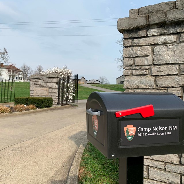 A black mailbox with a bright red flag and NPS arrowhead next to a stone pillared gate.