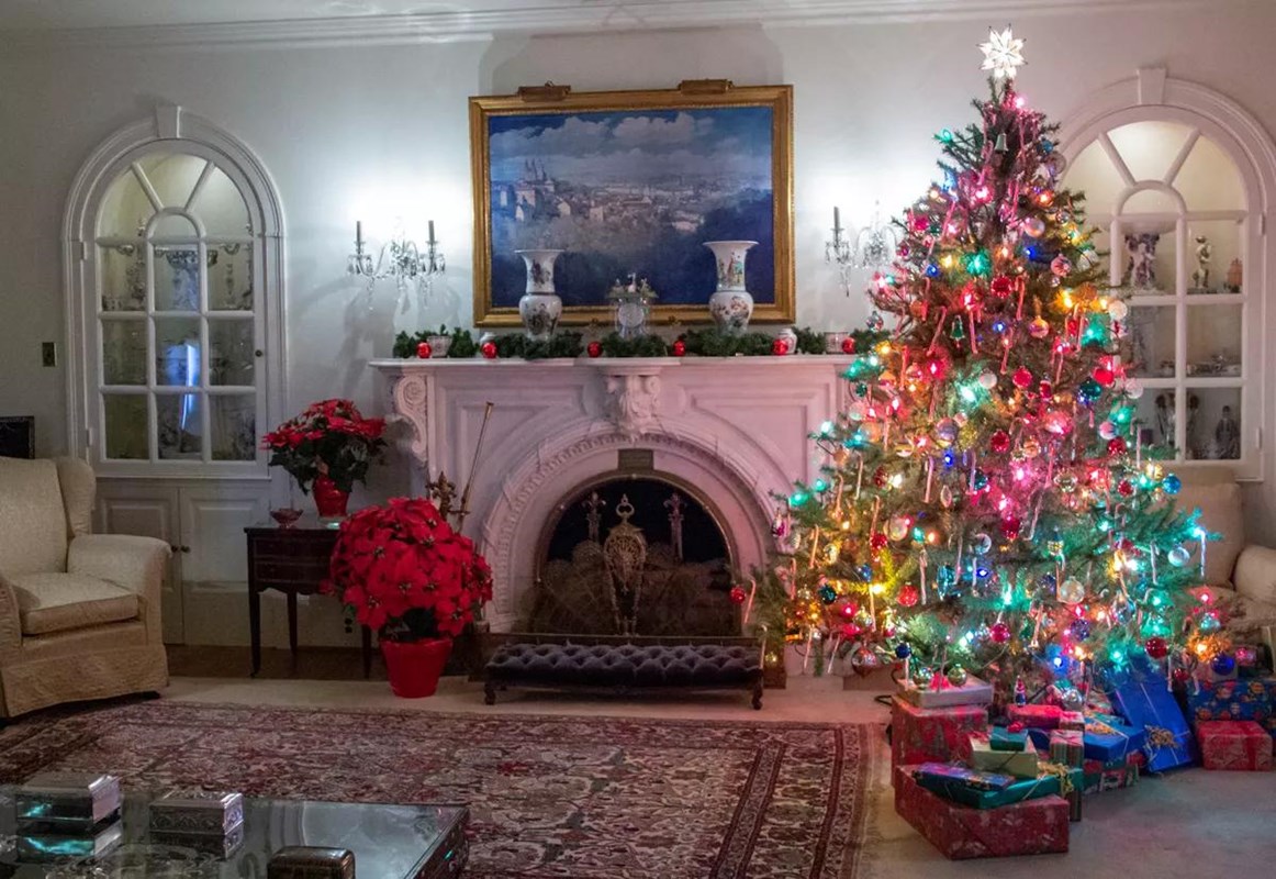 A color image of a Christmas tree next to a large rug and a white marble fireplace