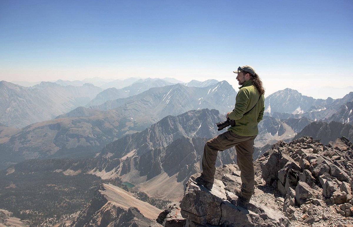 A man holding a camera standing atop a mountain, with other mountains in the distance.