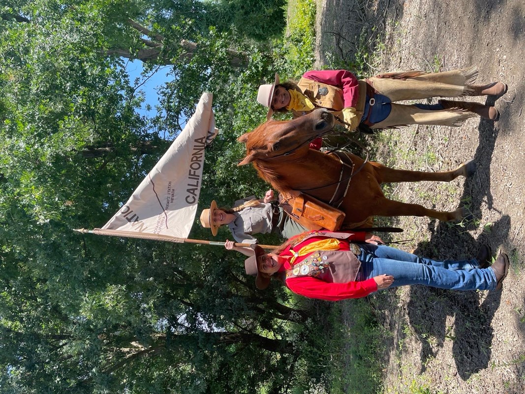 A park ranger sits on top of a horse holding a flag, while two people in western attire on the side.