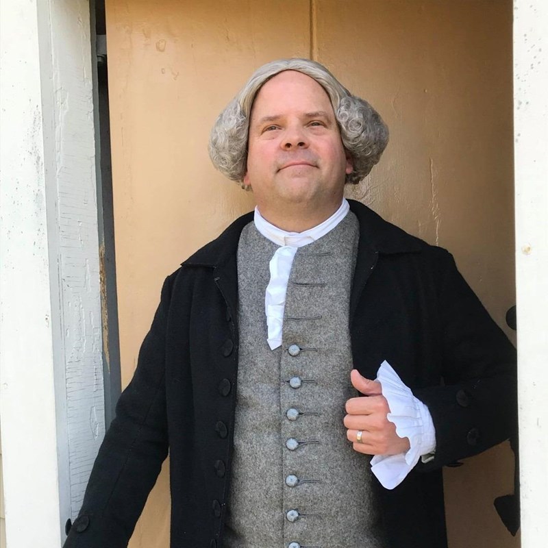 A man wearing 1700s-style wig and clothing stands in the door of a historic home