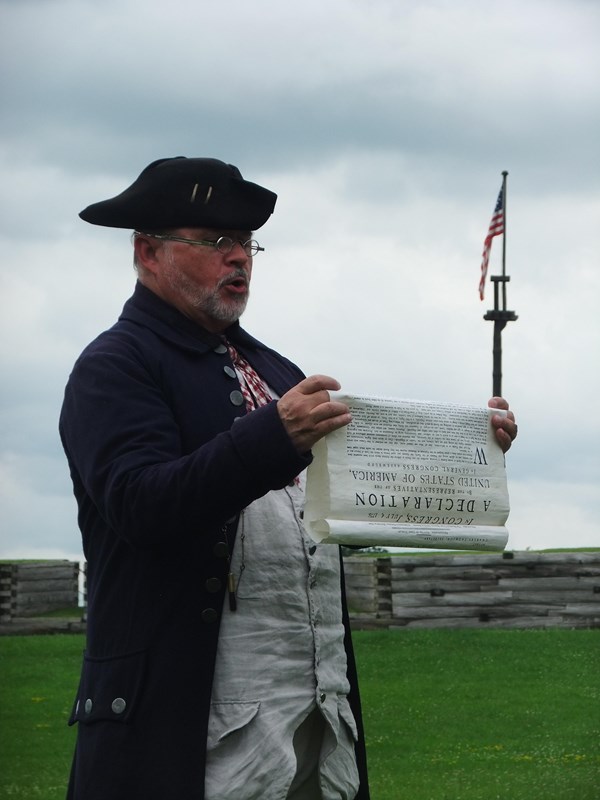 A man in 18th C garb stands in front of the fort while holding a large paper scroll.