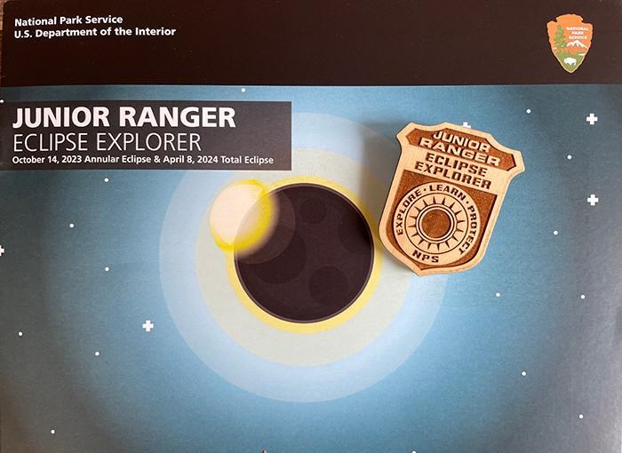 Cover of a booklet title Junior Ranger Eclipse Explorer with a badge on top of the booklet