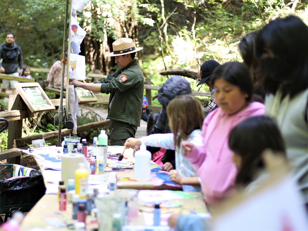 A ranger, left, posts art to a board with a table of supplies & children painting in the foreground