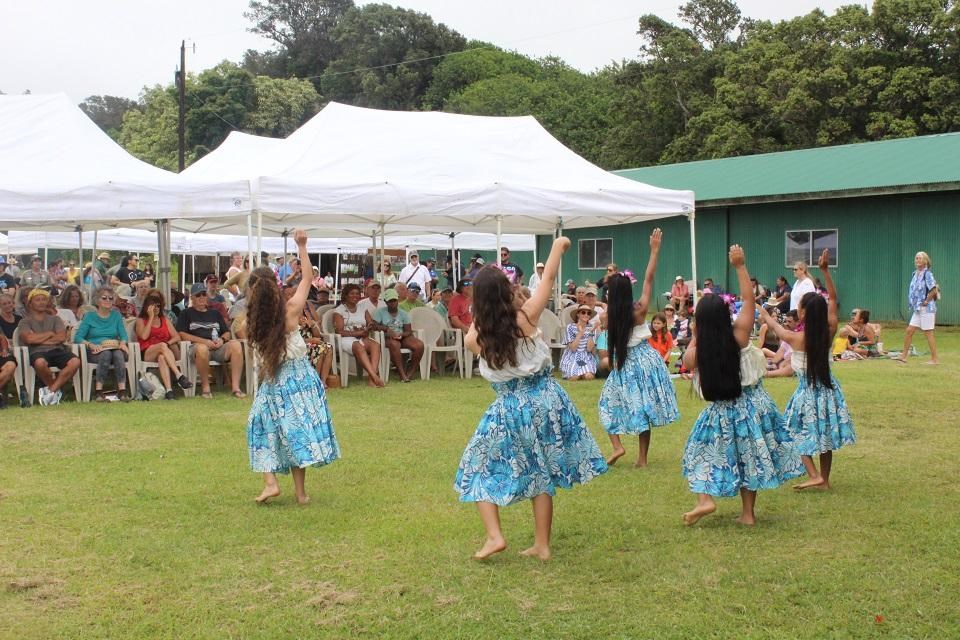 Young dancers perform hula in front of a seated audience on a lawn
