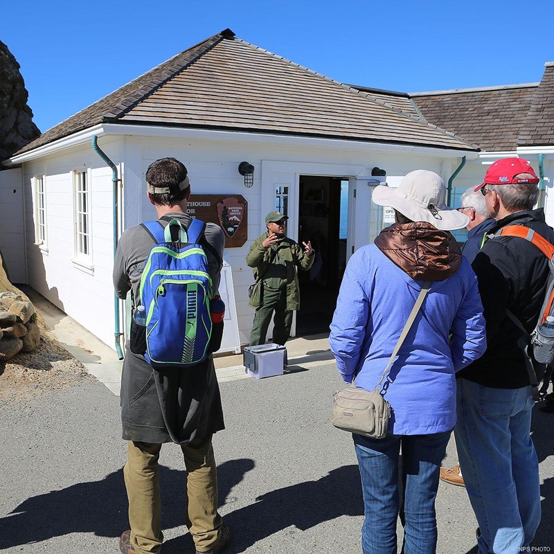 A park ranger dressed in green stands in front of a small white building talking to people.