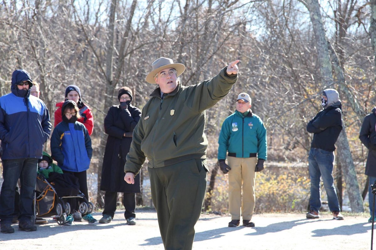 A park ranger standing with a crowd points to the landscape off camera.
