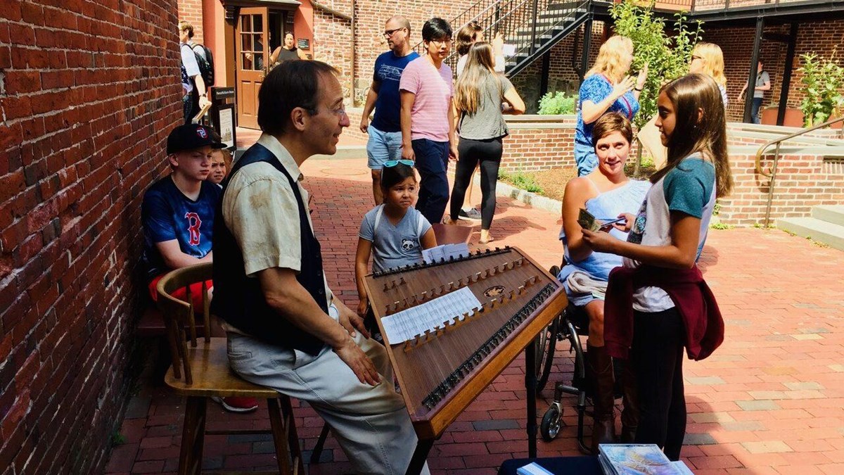Children crowd around a man in colonial cloths playing a Hammered Dulcimer