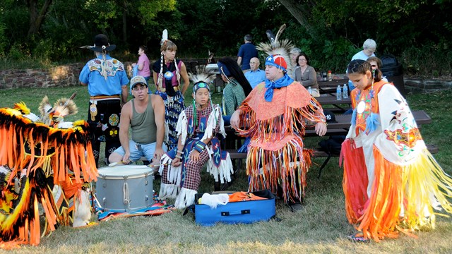 A group of Native Americans in ceremonial dress sit at a picnic table.