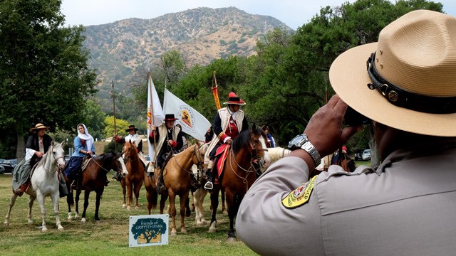 A park ranger in uniform takes a picture of a group of reenactors on horseback at a park.