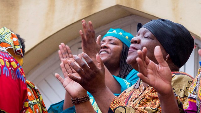 Two Black woman wearing colorful dresses and head wraps singing and clapping their hands