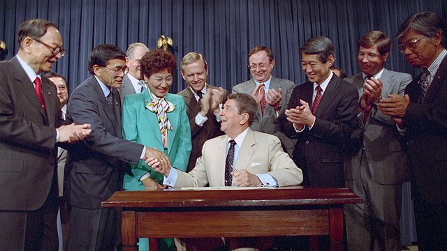 a group of people stand around a man seated at a desk clapping. one man shakes the seated man's hand