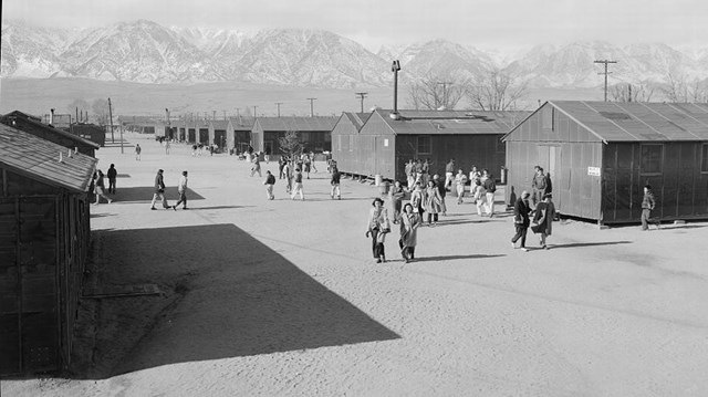 between rows of identical barracks people walk on a wide dirt street with mountain backdrop