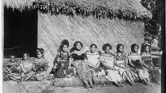 Black and white photograph of women and children of Tonga sitting in front of a straw building.