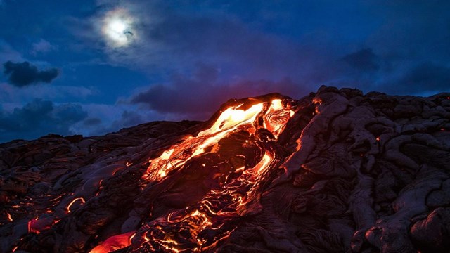 Molten lava spills out of a volcano during a cloudy night with the moon peeking through.